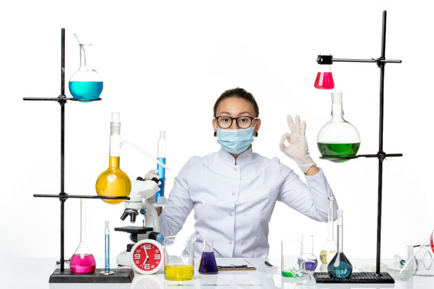 化学正面图：穿着医疗服的女化学家 戴着面罩 白色桌子上拿着装有蓝色溶液的烧瓶 飞溅着病毒化学实验室的病毒保持喷溅实验室