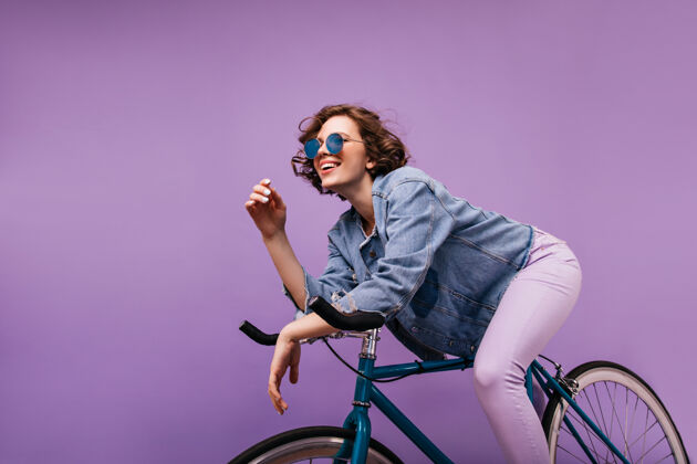 微笑无忧无虑的短发女士坐在自行车上快乐的高加索女孩 波浪式的发型表达积极的情绪爱好运动员紫色