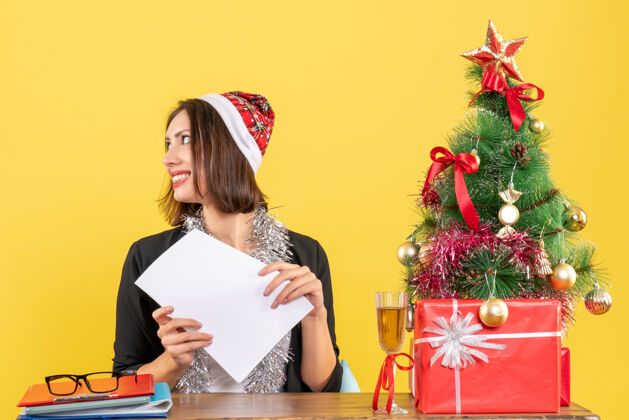 年一位身着西装 戴着圣诞老人帽 戴着新年装饰品 面带微笑的商务女士独自一人工作 坐在一张桌子旁 桌子上放着圣诞树独自人成人