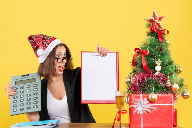 人迷茫迷人的女士穿着西装 戴着圣诞老人的帽子 展示着文件 手里拿着计算器 在黄色的办公室里与世隔绝西装帽子欢呼