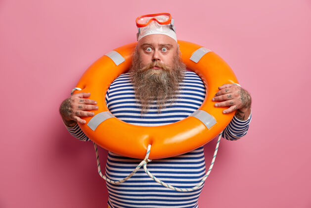 男人受惊的超重度假者害怕沉没 使用安全设备 戴潜水镜 用救生圈游泳 用震惊的表情直视旅游保险帽子监督穿着