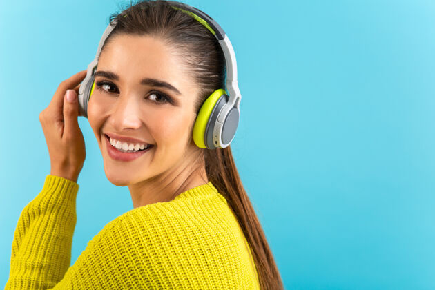 时尚迷人时尚的年轻女子戴着无线耳机听音乐快乐地穿着黄色针织毛衣五颜六色的时尚造型在蓝色背景上孤立地摆姿势微笑声音年轻