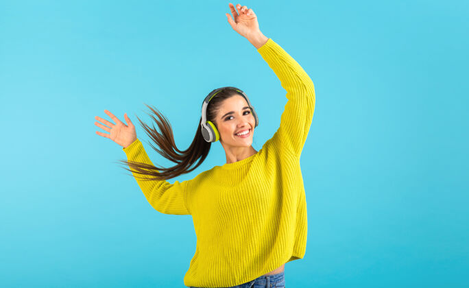 声音迷人时尚的年轻女子戴着无线耳机听音乐快乐地穿着黄色针织毛衣五颜六色的时尚造型隔离在蓝色背景上挥舞着长长的发尾立体声女孩音频