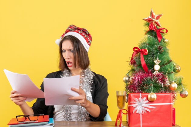 坐情绪混乱的商务女士穿着西装 戴着圣诞老人帽 戴着新年装饰品 看着头上的文件 坐在办公室里一张放着圣诞树的桌子旁树年西装