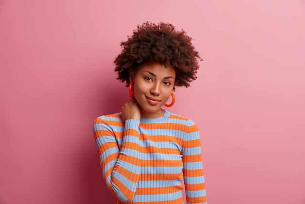 非洲可爱的黑皮肤千禧一代女孩的肖像摸着脖子 表情迷人 看到她想要的东西 卷曲浓密的头发 穿着随意 孤立在粉红色的墙上罗西毛衣条纹