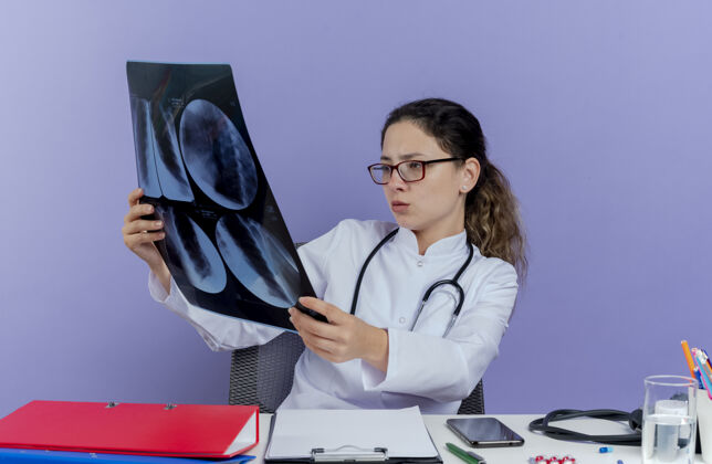 紫色专注的年轻女医生穿着医用长袍和听诊器坐在办公桌旁 手持医疗工具 看着x光片射击年轻人听诊器