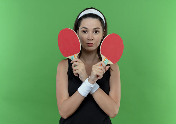 举行戴着头巾拿着球拍打乒乓球的年轻健身女正脸交叉双手站在绿色背景下看着摄像机相机头带立场