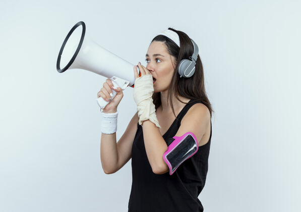 健身戴着耳机和智能手机袖标的年轻健身女士站在白色背景下对着扩音器大声喊叫手机年轻扩音器