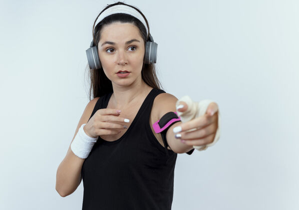 站立戴着头戴耳机和智能手机袖标的年轻健身女士站在白色背景上向外看手臂手机健身