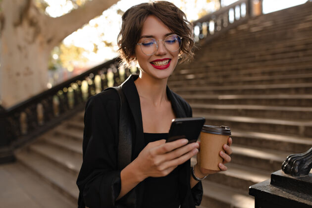 女性一个积极的女人 红唇 黑色套装 拿着电话和咖啡摆姿势戴着眼镜的卷发女人在外面微笑台阶步行阳光