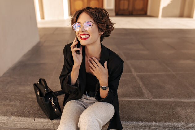 时尚戴着眼镜的黑发快乐的女人微笑着坐在外面穿着黑夹克白裤子的年轻女人在户外打电话路径外面台阶