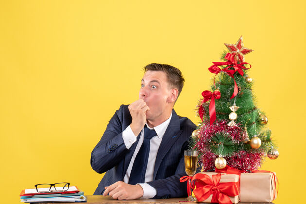 生意前视图令人惊讶的男子坐在圣诞树附近的桌子上 并提出了黄色背景上的自由空间坐着树西装