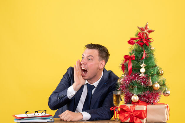 男前视图愤怒的男子一边大声喊叫 一边坐在圣诞树旁的桌子上 呈现黄色背景礼物前面人