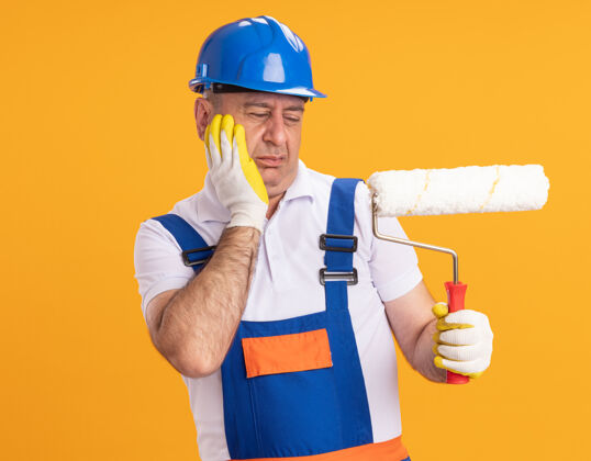 穿着穿着制服的未出租的成年建筑工人戴着防护手套 把手放在脸上 看着隔离在橙色墙上的滚筒刷手套滚轴手