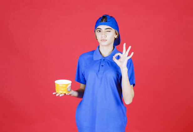 食物身着蓝色制服的女快递员手持黄色面杯 并展示满意标志生产力年轻人外卖