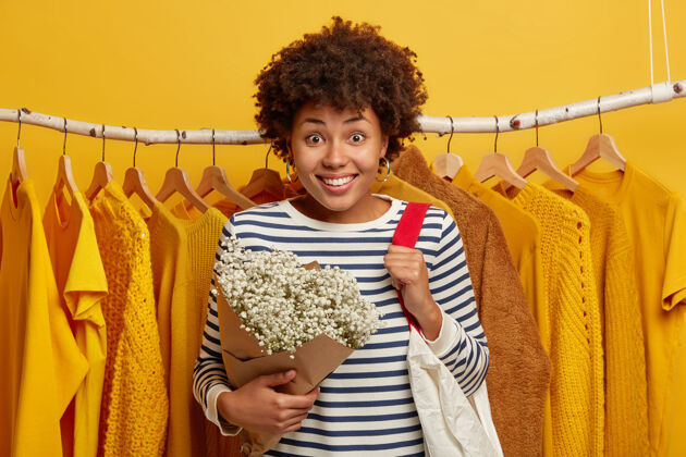 消费主义微笑的美国黑人女子对着衣柜摆姿势 选择合适的新装 喜欢黄色 拎包 捧花 笑容灿烂购买衬衫包