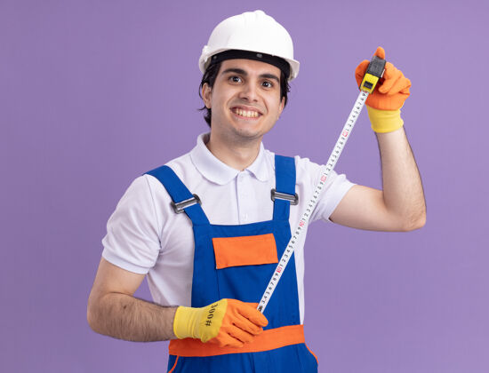 站着年轻的建筑工人穿着建筑制服 戴着安全帽 戴着橡胶手套 手里拿着卷尺 面带微笑地站在紫色的墙上看着前方安全建筑橡皮
