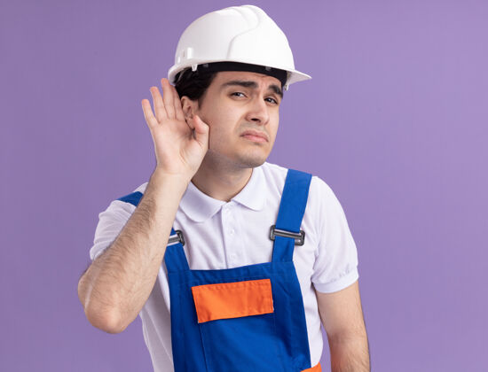 建筑年轻的建筑工人穿着建筑制服 戴着安全帽 用手捂着耳朵望着前方 试图倾听站在紫色墙壁上的流言蜚语头盔耳朵建筑工人