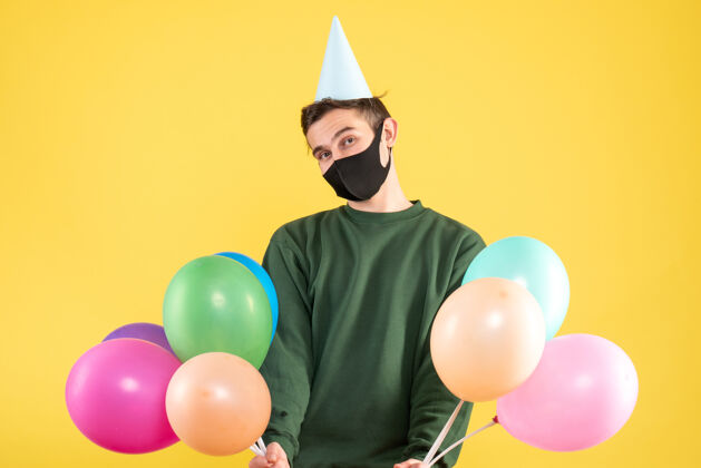 气球正面图：戴着派对帽和彩色气球的年轻人站在黄色的背景复制空间上帽子快乐年轻人