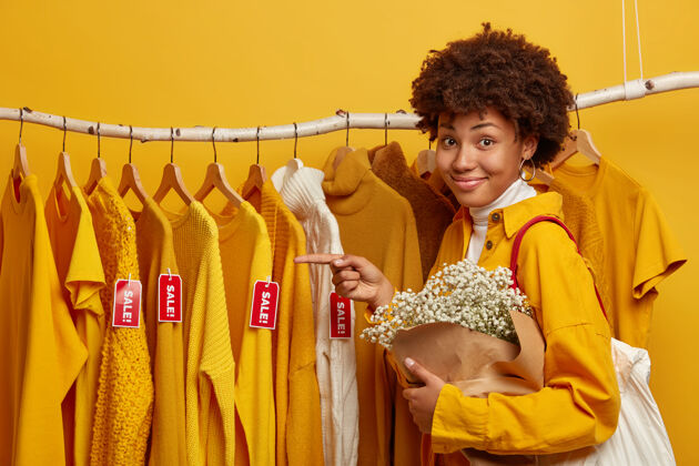 消费者友好的女顾客侧身站在衣架上 指着有标签的毛衣 肩上有购物袋 手里拿着花束女标签单色