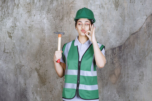 年轻戴绿色头盔的女工程师手持木柄斧头进行维修工作 并显示积极的手势幸运混凝土工具