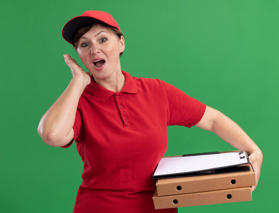 空白中年送货员身穿红色制服 头戴鸭舌帽 手里拿着披萨盒和写字板 手里拿着空白的书页 困惑而惊讶地站在绿色的墙边看着前面站着年龄帽子