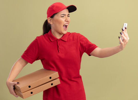 喊叫愤怒的年轻送货员身穿红色制服 戴着帽子 手里拿着比萨饼盒 看着她的智能手机 站在绿色的墙上 带着咄咄逼人的表情大喊大叫拿着愤怒咄咄逼人