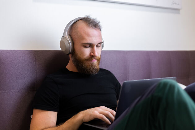 卧室一个留着大胡子的白种人戴着耳机在卧室的床上工作 在家里用笔记本电脑 打字 思考房间躺着技术