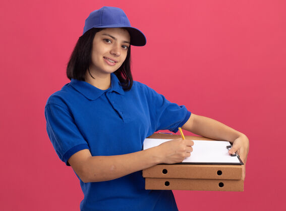 制服身穿蓝色制服 头戴鸭舌帽 手持比萨饼盒和剪贴板 面带微笑的小女孩站在粉红色的墙上拿着盒子站着