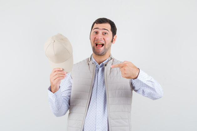 帅气年轻人指着他的帽子在衬衫 无袖夹克衫 看起来乐观前视图优雅肖像性感