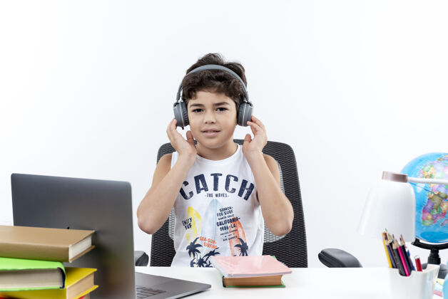 耳机看着镜头 戴着耳机的小男生坐在书桌旁 手里拿着学习工具 把耳机隔离在白色背景上小小学生手
