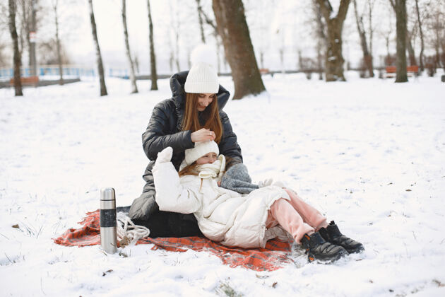 毯子妈妈和女儿坐在雪地公园的毯子上帽子孩子暖瓶