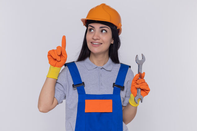 建筑工人身穿施工制服 头戴安全帽 戴橡胶手套 手持扳手 食指 微笑着站在白色墙壁上的年轻建筑工人安全显示橡胶