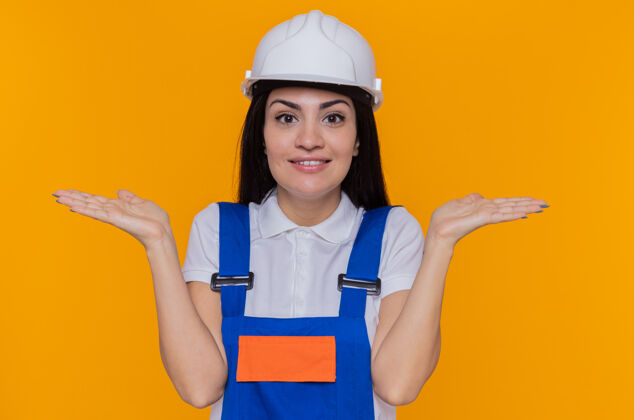 女人身穿施工制服 头戴安全帽的年轻建筑工人望着前方 面带微笑 兴高采烈地张开双臂站在橙色的墙上制服手臂微笑