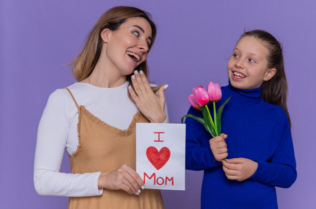 花快乐的女儿站在紫色的墙上 给她惊喜而微笑的母亲送上贺卡和郁金香花 庆祝国际妇女节微笑国际女人