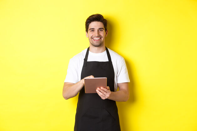 时尚帅气的服务员拿着点菜 拿着数码平板电脑 面带微笑 身穿黑色围裙制服 站在黄色的背景下咖啡师工作室自信