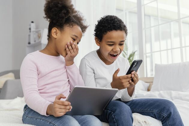 水平两个微笑的兄弟姐妹在家里一起玩平板电脑和智能手机手机房子室内