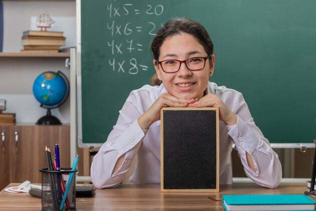 黑板年轻的女老师戴着眼镜拿着小黑板讲课看着前面笑容可掬地坐在课桌前教室里的黑板前眼镜坐着戴着