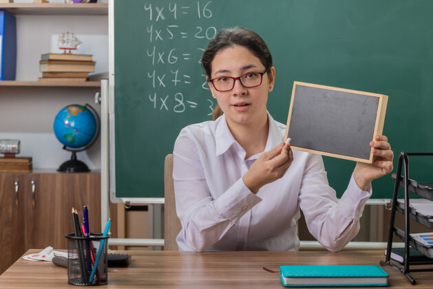 学校年轻的女老师戴着眼镜 手里拿着小黑板和粉笔讲解功课 面带微笑 自信地坐在教室黑板前的课桌旁解释粉笔女人