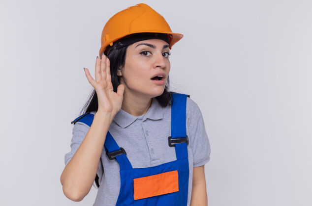 手一位身穿建筑制服 戴着安全帽的年轻建筑妇女 手捂着耳朵 试图倾听站在白墙上的流言蜚语年轻试着头盔