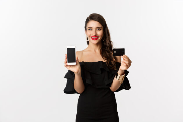 表情网上购物概念时尚的黑发女子穿着黑色连衣裙 展示手机屏幕和信用卡 微笑着高兴 站在白色背景下黑发信用卡美丽