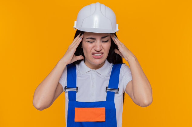 痛苦身穿施工制服 头戴安全帽的年轻建筑工人站在橘色的墙上 双手触碰头部 头痛得厉害女人年轻站立