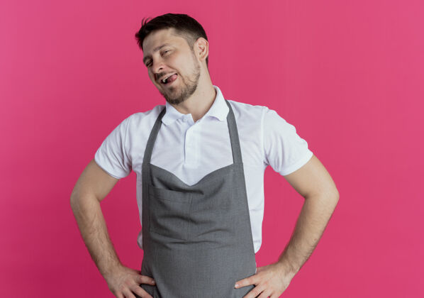 棍子围裙上的理发师伸出舌头 手臂放在臀部 站在粉红色的墙上男人理发师臀部