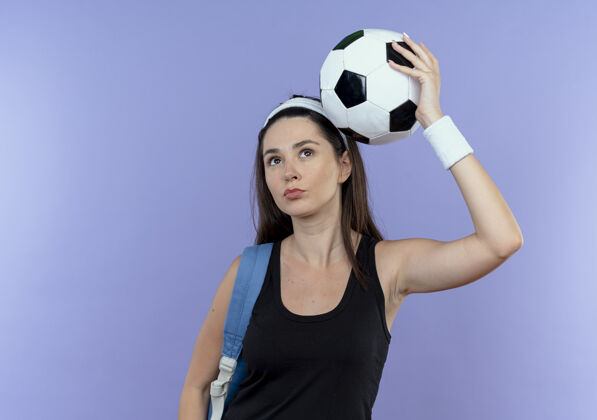 抱戴着头巾 背着背包 头上举着足球的年轻健身女士站在蓝色的墙上 看起来很自信健身头女子