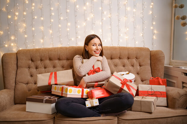 发光幸福的年轻女子抱着许多礼物盒盘腿坐在一个驼色沙发与灯圣诞节礼物拥抱