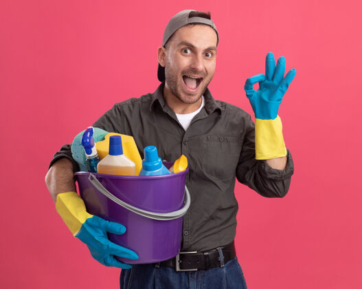 休闲年轻人穿着休闲服 戴着橡胶手套 戴着帽子 手里拿着带清洁工具的水桶 面带微笑 粉色的墙上挂着“ok”的牌子水桶男人站着