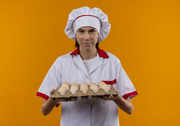 持有穿着厨师制服的年轻恼怒的白人厨师女孩拿着一批鸡蛋 看着被隔离在橙色空间和复印空间的摄像机厨师年轻看