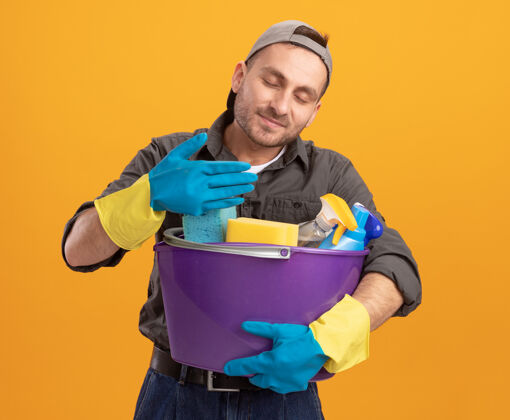 持有年轻人穿着休闲服 戴着橡胶手套 手里拿着带清洁工具的水桶 站在橙色的墙上 呼吸着清新宜人的香气衣服新鲜穿着