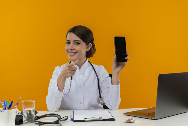 手机面带微笑的年轻女医生穿着医用长袍 听诊器坐在桌子旁 手持医疗工具和笔记本电脑 手机孤立地指向黄色的墙上听诊器微笑表演