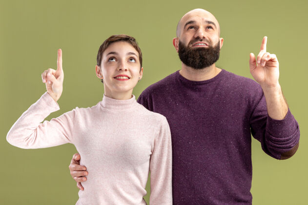 胡须一对穿着休闲服的年轻夫妇一位留着短头发和胡须的女士抬起头来 伸出食指 站在绿色的背景上 快乐而积极地庆祝情人节手指立场休闲